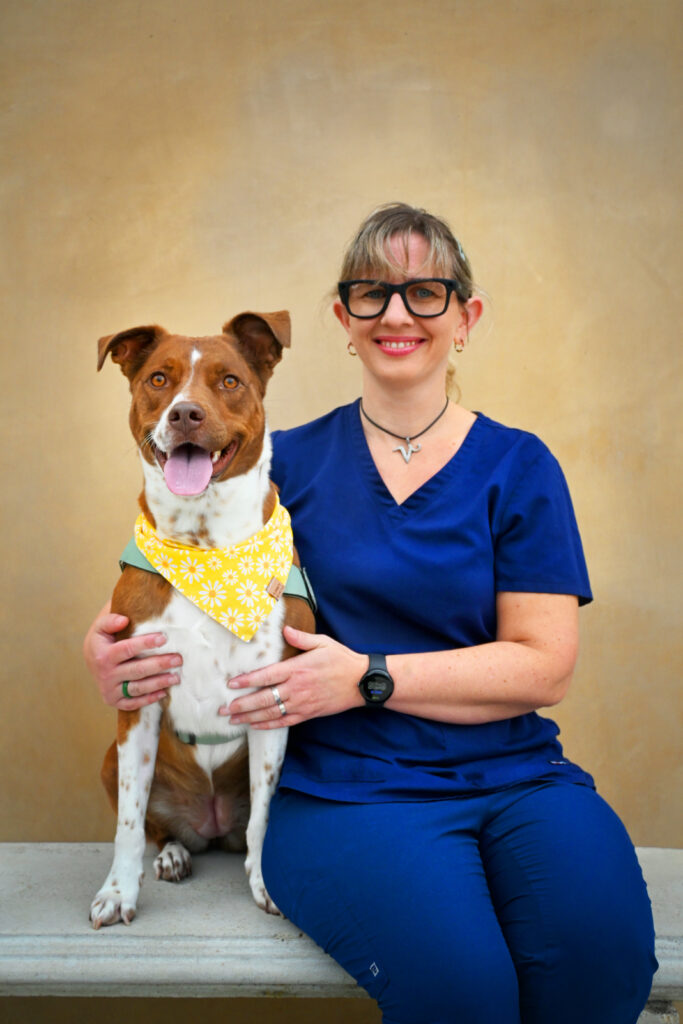 Blonde woman in blue scrubs and black glasses sitting with a brown and white dog on a bench.