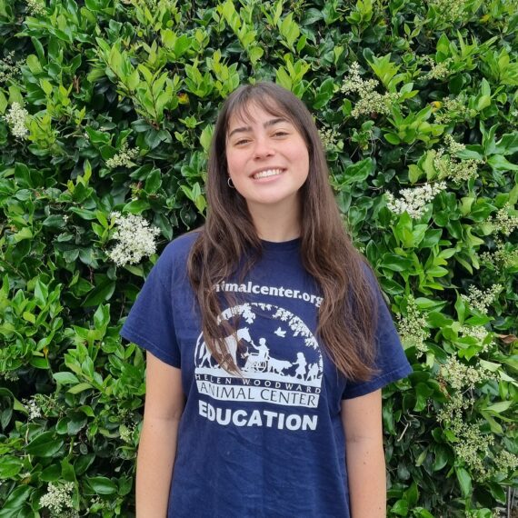 Young lady with long hair and a blue Helen Woodward Animal Center T-shirt standing in front of greenery